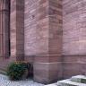 Teilansicht einer Kirchenwand aus rötlich grauem Mauerwerk mit Stützpfeilern und Eingangsbereich links hinten. Der Vorplatz ist gepflastert.