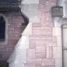 Teilansicht einer Kirchenfassade aus rötlich grauem Mauerwerk. Zu sehen ist rechts ein Vorbau mit Pfeiler und Türbogen; an den Rändern mit helleren Steinen abgesetzt. Links oben ist ein Ornament-Fenster erkennbar.