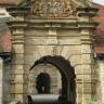 Blick auf das aus gelblich grauem Stein gefertigte äußere Tor eines Schlossbaues, mit tunnelartigem Durchgang, Giebelaufbau, Figuren und Wappentafel. Im Hintergrund ist ein weiterer Durchgang erkennbar.
