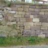 Das Foto zeigt eine Steinmauer mit gelblichen und rötlichen Steinen. Links verzahnt sich die Mauer mit einer unregelmäßigen Steinbruchwand.