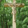 Nahaufnahme eines Friedhofskreuzes mit Christusfigur aus gelblich grauem Stein. Im Hintergrund stehen hohe, dicht belaubte Bäume.