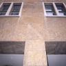 Aufwärts gerichteter Blick auf eine Gebäudefassade aus gelblich grauen Steinplatten. Unten teilt eine Säule die Fassade, darüber verlaufen Fensterreihen.