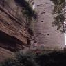 Blick auf eine hohe, rötlich braune Gesteinswand (links im Bild) sowie einen daran angebauten runden Turm aus rötlich grauem Mauerwerk (rechts). Kuppe und Fuß der Steinwand sowie das Fundament des Turmes sind von Sträuchern bewachsen.