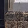 Teilansicht eines Gebäudesockels aus gelblich bis rötlich grauen, stark schraffierten Mauersteinen. An die Mauerkrone schließen sich grüngraue Türrahmen sowie eine blaugraue Randleiste aus glattem Gestein an. 