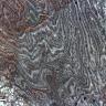 Detailaufnahme einer Gesteinsoberfläche mit welliger bis streifiger Fließstruktur, im Wechsel von grauem und weißem Material. Auf der linken Seite des Bildes sind auch braune Farben erkennbar.