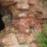 Teilansicht einer gelblich braunen Steinbruchwand, mit rötlichen hervortretenden Gesteinskörpern. Links ist ein Hammer angelehnt.