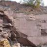 Blick auf eine rötlich graue Steinbruchwand mit großen Blöcken rechts und zerklüftetem Gestein links im Bild. 