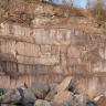 Blick auf eine hohe, oben bewachsene Steinbruchwand, teilweise mit senkrecht verlaufenden Spuren von Abbauarbeiten. Im Vordergrund liegen Bruchstücke auf einem Haufen. 