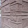 Teilansicht eines von einem rechteckigen Pfeiler gestützten Gebäudes aus braunrotem Gestein, mit Ecksteinen, dünnen Zwischenplatten und Quadern mit unterschiedlicher Oberfläche. 