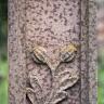 Teilansicht eines rötlich grauen, steinernen Grabkreuzes mit herausgearbeiteter Verzierung in Form einer Blume mit zwei Kelchen und Blättern. Der Stein weist zudem zahlreiche dunklere Flecken auf.