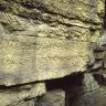 Seitlicher Blick auf eine Steinbruchwand mit grünlich grauem Gestein und zahlreichen tiefen Einkerbungen. Im unteren Teil des Bruchs ist zudem eine Nische erkennbar.