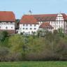 Blick auf eine größere Klosteranlage mit Hauptgebäude rechts und Nebengebäuden in der Mitte und links. Die Häuser sind weiß, mit braunen Ecken und Fensterrahmen. Das Gebäude links hat zudem einen braunen Steinsockel sowie Fachwerk. Die Dächer sind rot.