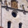 Teilansicht einer Kirchenfassade aus grauem bis hellbraunem Mauerwerk. Zu sehen sind Fenster mit runden Bögen, ein Rundfenster, Figuren und ein bronzefarbenes Relief oberhalb der Bildmitte.
