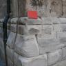 Teilansicht eines Gebäudesockels aus großen grauen, teils abgerundeten Mauerquadern. Ein zwischen Sockel und anschließendem Mauerwerk aufgelegtes rotes Buch dient als Größenvergleich.