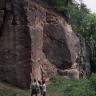 Blick auf eine massige, dunkle Felswand, die sich vor einem Waldhang befindet. Links durchzieht eine senkrechte Kluft das Gestein, rechts der Bildmitte ist eine größere Nische im Fels. Zwei Personen stehen am Fuß der Felsformation.