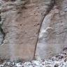 Teilansicht einer bräunlichen bis grauen Gesteinswand. Über einem glatten, etwas abgesetzten Sockel ist das Gestein gefurcht und löchrig. Am Fuß liegt von Schnee bedecktes Laub.