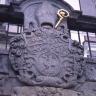 Nahaufnahme eines aus violettgrauem Stein gearbeiteten Wappens. Im oberen Teil ist eine Bischofsmütze sowie ein vergoldeter Stab erkennbar. Das Wappen ist Teil einer Gebäudefassade oder einer davor angebrachten Mauer.