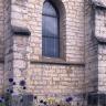 Teilansicht einer Kirchenfassade aus bräunlich grauem Mauerwerk mit beidseitigen Stützpfeilern und mittig platziertem Spitzbogenfenster.