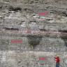 Zu sehen ist eine graue Felswand, die sich horizontal durch unterschiedliche Grautöne gliedert. Die einzelnen Schichten sind durch gestrichelte Linien und Beschriftungen unterschieden. Am Fuß der Felswand steht eine Person mit Schutzhelm und Maßstab.