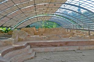 Blick auf die Überreste eines antiken römischen Bades. Im Vordergrund der Einstiegsbereich mit Treppenaufgang links, dahinter gemauerte Einfassungen und Ziegelsteinwände. Geschützt werden die Ruinen von einem halbkreisförmigen Gitterdach aus Metall.