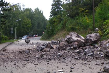 Das Bild zeigt eine Straße, auf die von einem Hang rechts Steine und Felsbrocken geschoben wurden. Die Straße ist abgesperrt. Ein Mann links in Warnkleidung begutachtet den Schaden.