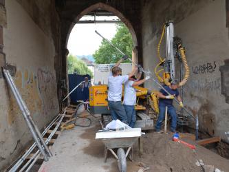 Das Bild zeigt die Baumaßnahmen zur Sanierung des Schwabentors. Drei Männer führen ein Bündel dünner Metallrohre schräg in den Boden ein. Vor den Männern steht eine Schubkarre, dahinter ein Kleinbohrgerät. Ein weiterer Mitarbeiter trägt Metallrohre. 