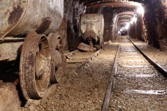 Besucherbergwerk Tiefer Stollen – Blick in die Tiefe Strecke; Foto: Stadt Aalen/Besucherbergwerk Tiefer Stollen Blick aus der Hocke auf Gleise und Beförderungswagen eines Bergwerkes. Rechts führen die Gleise geradeaus in einen beleuchteten Schacht.