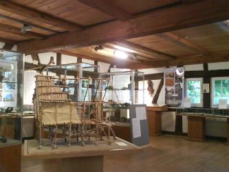 Blick in das Innere eines Museums mit Schautafeln neben den Fenstern, Schaukästen und Glasvitrinen. Links der Mitte ist das Holzmodell eines Pfahlbauhauses aufgestellt.