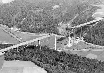 Das schwarzweiße Luftbild zeigt den Bau einer Brücke, über die eine mehrspurige Straße führen soll. Die Brücke überspannt ein Flusstal.