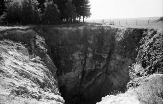 Altes Schwarzweißfoto, das den tiefen Krater einer Doline zeigt. Das freiliegende Boden- und Gesteinsmaterial befindet sich auf einer Wiese am Rande eines Waldes.
