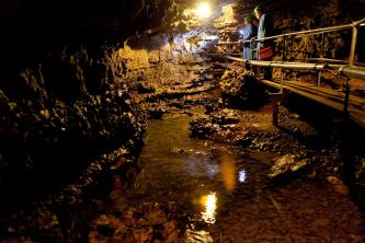 Blick in eine mit Lampen ausgeleuchtete Höhle, deren Boden mit Wasser bedeckt ist. Rechts verläuft ein schmaler Laufgang mit beidseitigem Geländer.