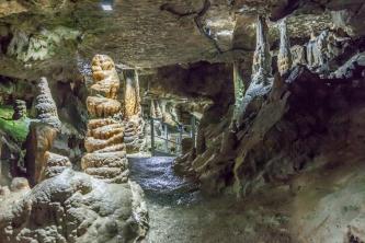 Blick in eine gut ausgeleuchtete Höhlenkammer mit Tropfsteinsäulen. Die Decke im Vordergrund ist sehr niedrig. Ein Laufgang in der Bildmitte führt in eine weitere, tiefer liegende Kammer.