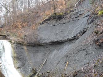 Blick auf einen grauen, oben links bewaldeten Gesteinshang mit bogenförmiger Verwerfung in der Bildmitte. Links unten ist ein Wasserfall angeschnitten.