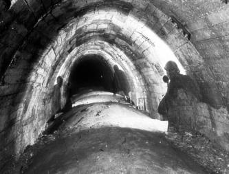 Ältere Aufnahme in Schwarzweiß von einem schmalen, nach oben runden Tunnel. Der Tunnelboden ist leicht nach oben gewölbt, die Tunnelwände sind gemauert. Auf beiden Seiten sind die Schatten des Fotografen zu erkennen.
