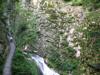 Rechts sieht man eine Felswand, die stellenweise von Gräsern und Sträuchern überwachsen ist. In der Mitte fließt ein steiler Fluss. Am linken Hang befindet sich eine lange Treppe.