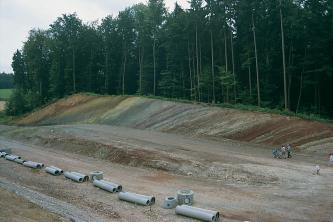 Blick auf eine Straßenbaustelle mit Röhren und Verbindungsstücken im Vordergrund, einer nach links an- und wieder absteigenden Rampe und einem angeschnittenen, bewaldeten Hang im Hintergrund. Der Hang weist schräg verlaufende, unterschiedliche Farben auf.