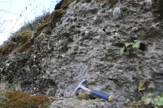 Blick auf einen nach rechts ansteigenden, grauen Felshang mit verbackenen und weggebrochenen Steinen. Links ist der Hang teilweise mit braunem Moos bewachsen. Rechts unten wurde ein Hammer angelehnt.
