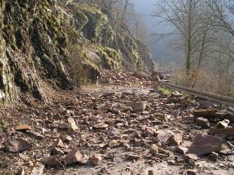 Blick auf eine Straße, die an einem links aufragenden Felshang entlangführt. Die Straße ist mit Steinen und Felstrümmern übersät und somit gerade unbefahrbar.