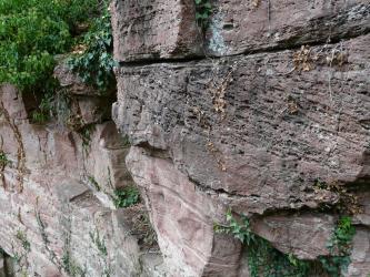 Seitlicher und leicht abwärts gerichteter Blick auf rötlich graue, rechts stark zerfurchte Steinblöcke, die zu einer Mauer zusammengefügt wurden. Einzelne Ritzen sowie die Mauerkuppe sind bewachsen.