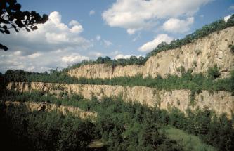 Blick auf drei in leichtem Bogen verlaufende Sohlen eines alten Steinbruchs. Die oberste Sohle steigt nach rechts hin an. Das helle Gestein des Bruchs ist jeweils an den Kuppen sowie am Fuß der Sohlen bewachsen.