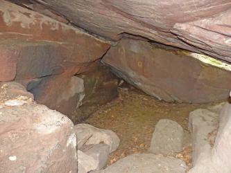 Blick in eine Felsenkammer. Die Kammer besteht aus rötlichen und grauen Felsbänken, die über Eck und schräg geschichtet stehen. In der Bildmitte ist zudem eine kleine Höhlenöffnung erkennbar.