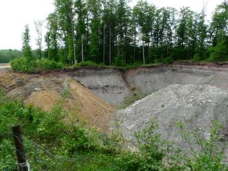 Gipsbruch am Rand des Reußenbergs südlich von Triensbach Das Bild zeigt die trichterförmigen Wände eines Steinbruches. Das Gestein, an einem Waldrand gelegen, ist grau mit rötlichen Stellen. Im Vordergrund sind zwei Abraumhalden aufgehäuft, die linke ist orangefarben, die rechte grau.
