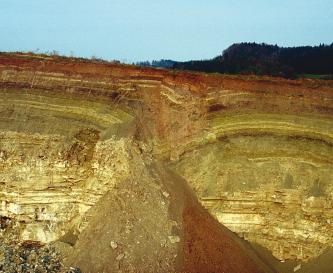Grabfeld-Formation (kmGr, früher: Gipskeuper) mit Doline: Steinbruch westlich von Gaildorf-Eutendorf Blick auf mehrere Schichten einer Steinbruchwand, die oben mit rötlichen Tönen beginnen, dann grünlich und schließlich gelblich werden. In der Mitte ist ein Einsturz zu erkennen, rechts bogenförmige Streifen.
