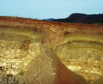 Blick auf mehrere Schichten einer Steinbruchwand, die oben mit rötlichen Tönen beginnen, dann grünlich und schließlich gelblich werden. In der Mitte ist ein Einsturz zu erkennen, rechts bogenförmige Streifen.