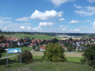 Blick über eine hügelige Landschaft. Im zum Hintergrund hin ansteigenden, flacheren Mittelgrund befindet sich eine Ortschaft. Am Horizont sind Waldstreifen erkennbar.