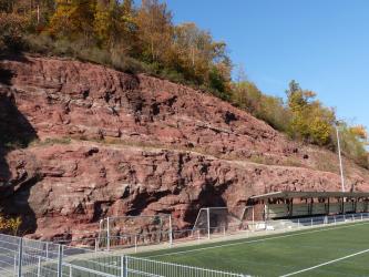 Direkt hinter einer Sportanlage (rechts, im Vordergrund) erhebt sich eine mächtige, abgestufte Gesteinswand. Das Gestein ist rötlich grau und steigt nach links hin an. Die Kuppe ist bewaldet.