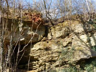 Blick auf zwei Wandseiten eines alten Steinbruchs, die in der Bildmitte - leicht verzahnt - aneinanderlehnen. Die rechte Wandseite steht dabei im Sonnenlicht; sie ist grünlich grau und teils zerfurcht. Links unten ist eine Nische oder Höhle erkennbar.