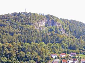 Blick auf einen stark bewaldeten, steilen Berg. Oberhalb der Bildmitte sind felsige Lücken im Berg erkennbar. Rechts unten, am Fuß des Berges, stehen Häuser.