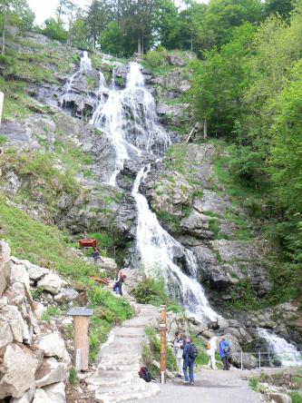 Blick auf eine hohe, abgestufte Felswand, über die ein breiter Wasserfall stürzt. Auf einem Treppenweg links steigen Besucher hinauf. Rechts wachsen Bäume am Felshang.