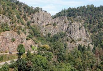 Die Südwestseite des Falkensteins südlich von Berau Blick auf mehrere große, rötlich graue Felsformationen, die aus einem steilen Waldhang hervortreten. Links, am unteren Rand einer hohen Felswand, windet sich eine Straße in spitzen Kehren abwärts.